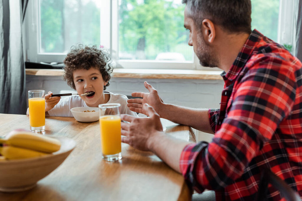 foco seletivo do menino encaracolado comendo flocos de milho e olhando para o pai perto de tigela e copos de suco de laranja
  - Foto, Imagem