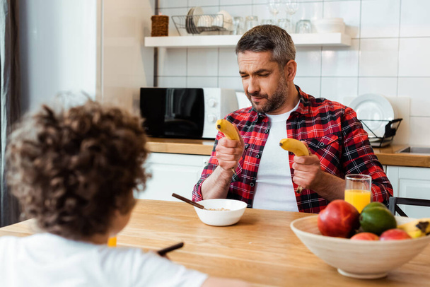selective focus of father holding fresh bananas near curly boy and breakfast on table  - Φωτογραφία, εικόνα