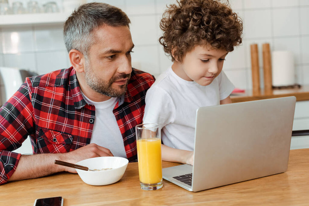 handsome father and curly son looking at laptop in kitchen  - Foto, Imagen