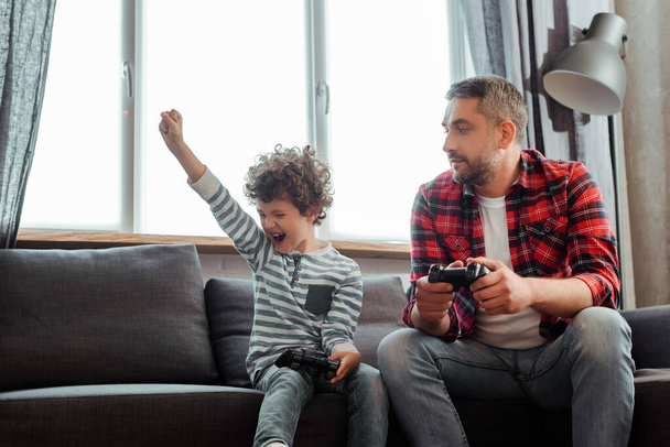 KYIV, UKRAINE - MAY 14, 2020: handsome father looking at excited curly son celebrating triumph while playing video game in living room  - Foto, Imagem