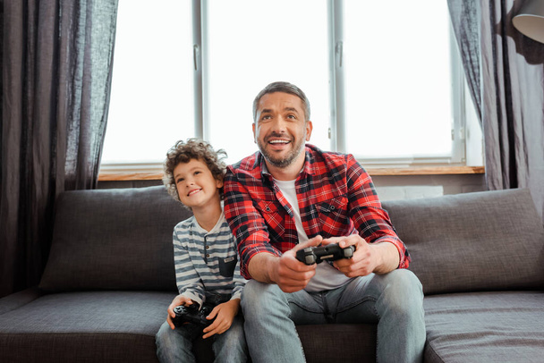 KYIV, UCRANIA - 14 de mayo de 2020: padre guapo e hijo feliz jugando videojuegos en la sala de estar
  - Foto, Imagen