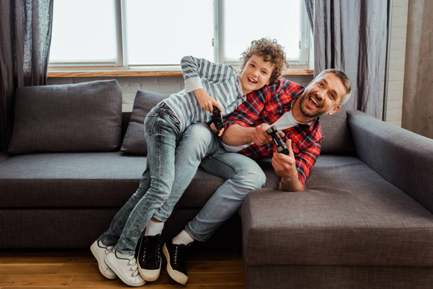 KYIV, UKRAINE - MAY 14, 2020: happy father and cheerful son playing video game in living room  - Foto, Imagem