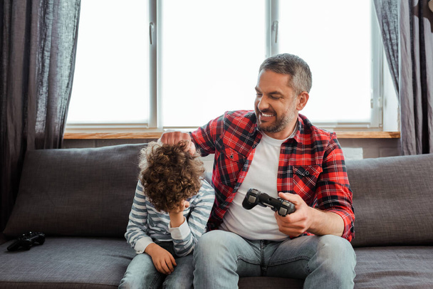 KYIV, UKRAINE - MAY 14, 2020: happy father touching upset and curly son  while playing video game in living room  - Φωτογραφία, εικόνα