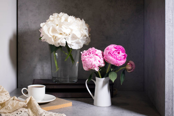 White hydrangea and peony in a glass vase with a small cup of coffee on a book with a notebook on a gray craft background. . Copy space - Фото, изображение