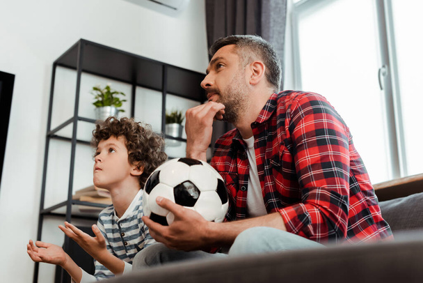 selective focus of bearded father holding football and watching championship with curly son - Foto, Bild