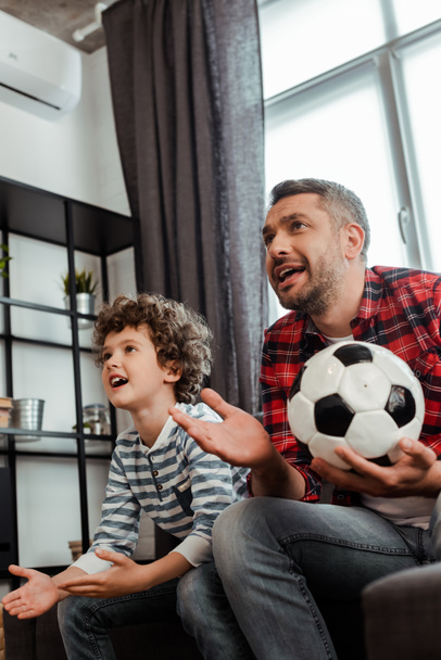 emotional and bearded father holding football and watching championship with curly son - 写真・画像