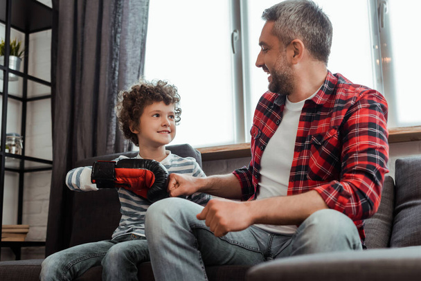 happy kid in boxing glove bumping fists with cheerful father in living room - Foto, Imagen