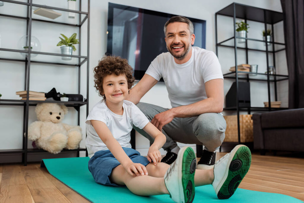 cheerful man smiling near happy son on fitness mat  - Photo, Image