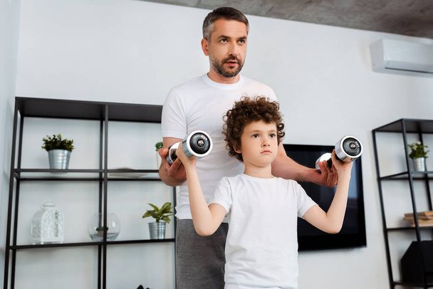 handsome father helping curly son exercising with dumbbells  - Photo, Image