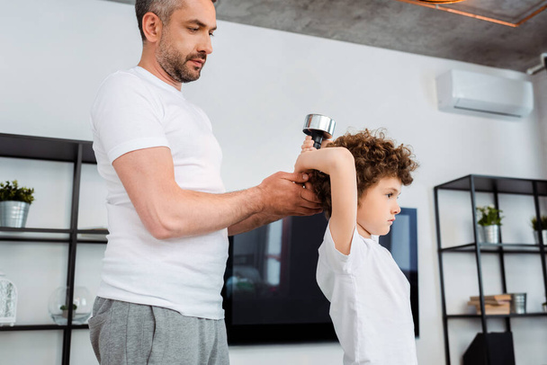 bearded father touching dumbbell while curly son exercising at home - Valokuva, kuva
