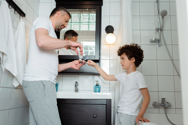 handsome father holding toothpaste near toothbrush of curly son  - Zdjęcie, obraz