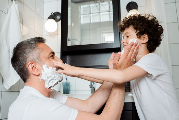 side view of happy father and cute son with shaving foam touching faces in bathroom  - Foto, immagini
