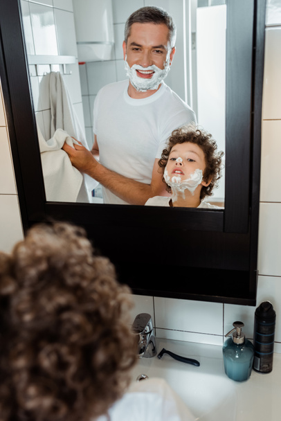 selektivní zaměření kudrnatého chlapce s pěnou na holení na obličeji při pohledu do zrcadla poblíž otce  - Fotografie, Obrázek