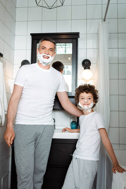happy father and curly son with shaving foam on faces looking at camera in bathroom - Photo, image