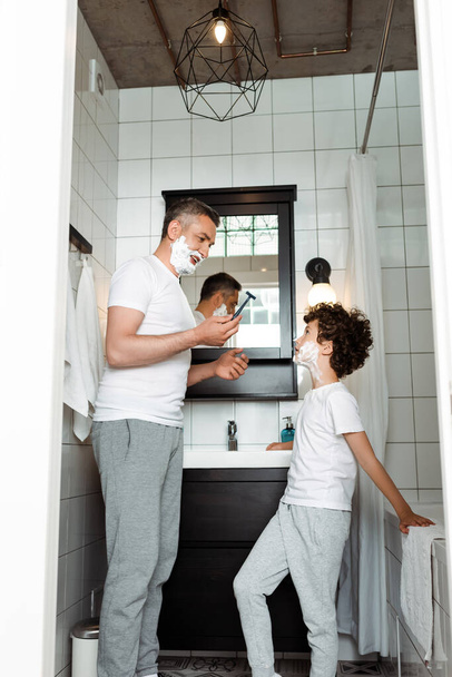 handsome father with shaving foam on face holding razor near curly son  - Foto, Imagem