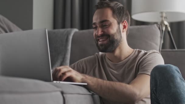 A pleased smiling young man is using his laptop sitting at home in the living room - Metraje, vídeo