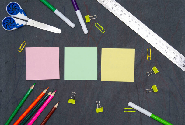 on a blackboard are scissors and a ruler, plasticine and colored pencils, paper clips and stickers in center. primary school education. flat lay. copy space. - Photo, Image