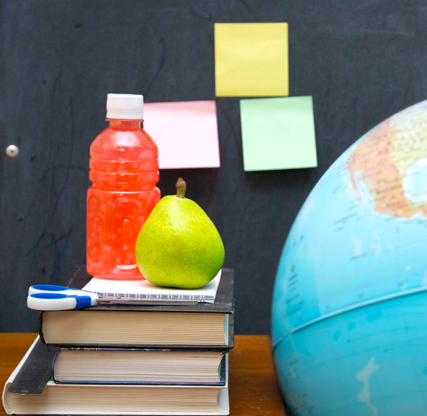juice and pear on top of a stack of textbooks on a desk against the background of a school board with sticks and a globe. concept school, education, lunch break. - Photo, Image