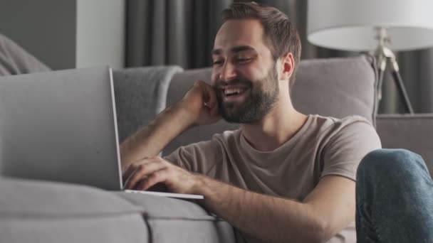 Um jovem feliz está digitando em seu laptop sentado em casa na sala de estar
 - Filmagem, Vídeo