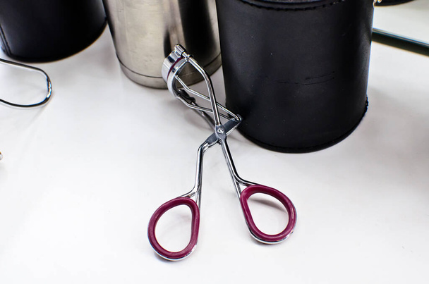 Stainless steel eyelash curler on white table with silver and black metal jars for brushes  - Photo, Image