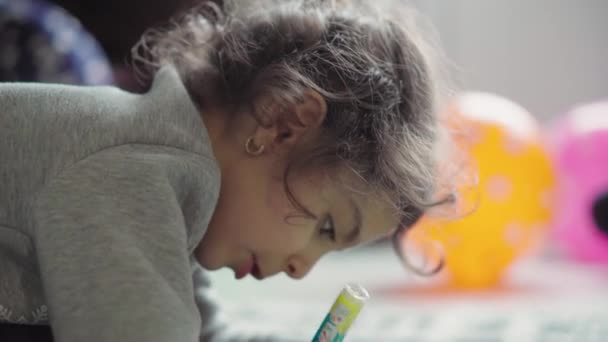 Childhood, play, training, self-isolation concept - close-up dark-haired curly little six-year-old girl of Persian Middle Eastern appearance draws in notebook pencil lying on her stomach on floor. - Video, Çekim