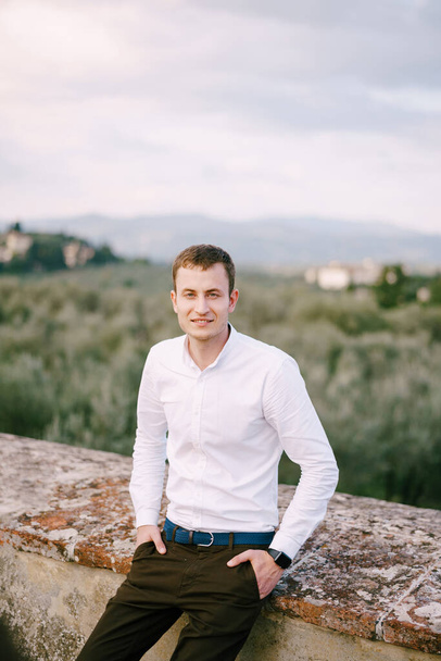 Een groot mannelijk portret op het dak van een oude wijnmakerij villa in Toscane, Italië. Wit shirt met lange mouwen, slimme horloges, blauwe riem, handen in broekzakken. - Foto, afbeelding