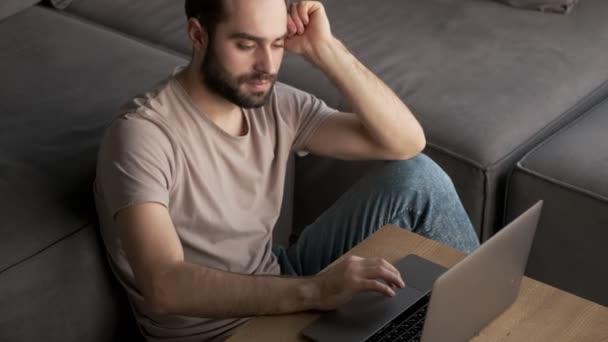 A stylish positive young man is using his laptop computer while sitting at home in the living room - Πλάνα, βίντεο