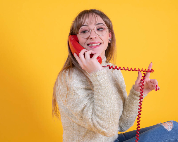 egy gyönyörű fiatal nő szemüveges egy régi piros telefon mosolygós kifejezés, sárga háttér - Fotó, kép
