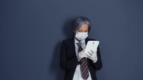 Grey haired businesswoman in protective mask and gloves holding and looking at tablet standing in the dark blue wall background . Health care concept. Business concept. Prores 422 - Filmagem, Vídeo