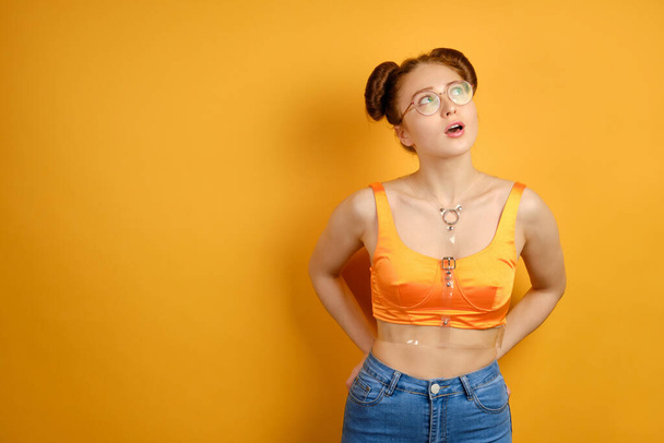 The girl in an orange top and glasses looks to the top, opening her mouth and putting her hands in the back pockets of jeans - Foto, Imagen