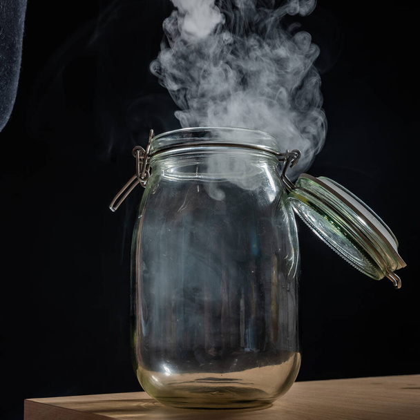 Glass jar with a lid filled with smoke on a black background. Fog in a transparent container on the table. - Foto, Bild