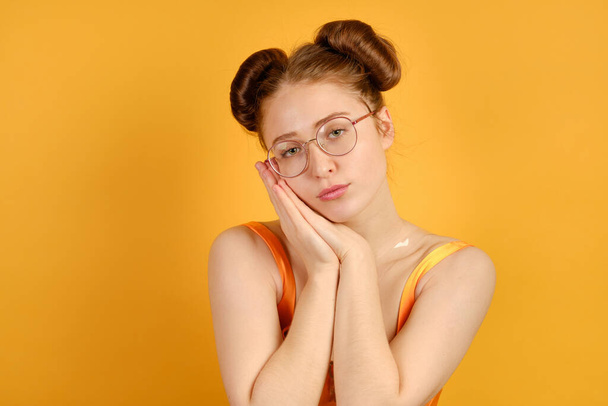 A red-haired girl in an orange top and round glasses, looks tiredly at the camera, resting her head on folded palms - 写真・画像