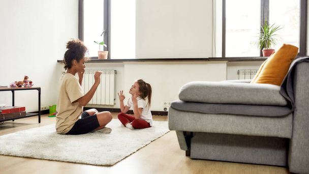 Healthcare. African american woman baby sitter and caucasian cute little girl doing yoga at home - Valokuva, kuva