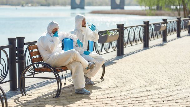 Hard workers. Sanitization, cleaning and disinfection of the city due to the emergence of the Covid19 virus. Specialized team in protective suits and masks takes a break near the riverside - Zdjęcie, obraz
