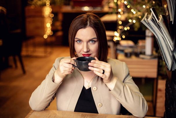 una joven con el pelo oscuro está sentada en un café y sosteniendo una taza de café o té en dos manos
 - Foto, Imagen