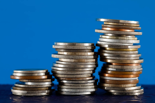 Διάφορα κέρματα τοποθετημένα σε τρεις στοίβες και απεικονίζονται σε μπλε φόντο. Η επιφάνεια στην οποία τοποθετούνται αυτά τα κέρματα είναι σκούρο μπλε. - Φωτογραφία, εικόνα