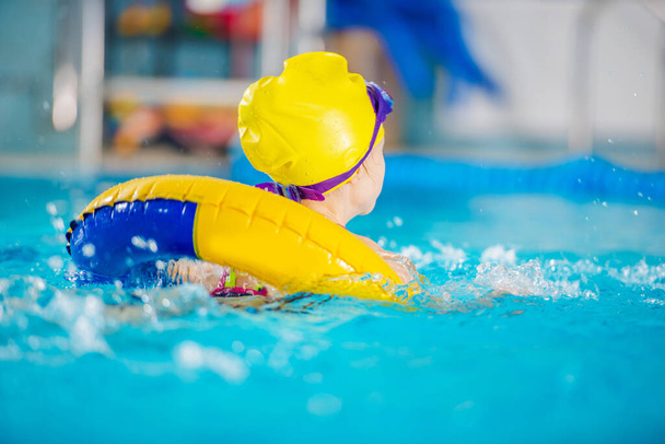 Watersport en Recreatie Thema. Jong meisje zwemmen in het grote overdekte zwembad Binnen gele opblaasbare buis. Draag een badmuts en een bril. - Foto, afbeelding