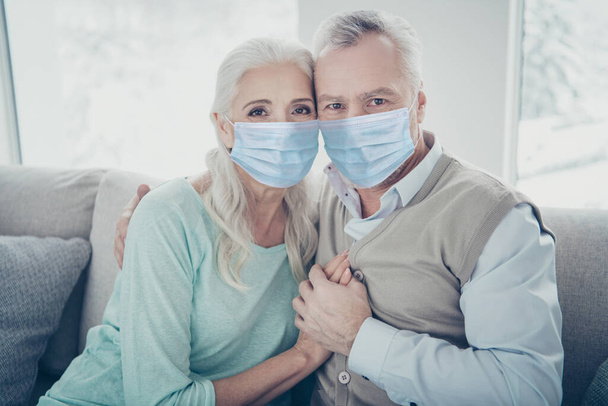 Photo of pretty elderly aged couple bonding leaning heads holding hands sitting on cozy sofa wearing gauze mask stay home preventive measure risk social group home house flat apartment - Zdjęcie, obraz