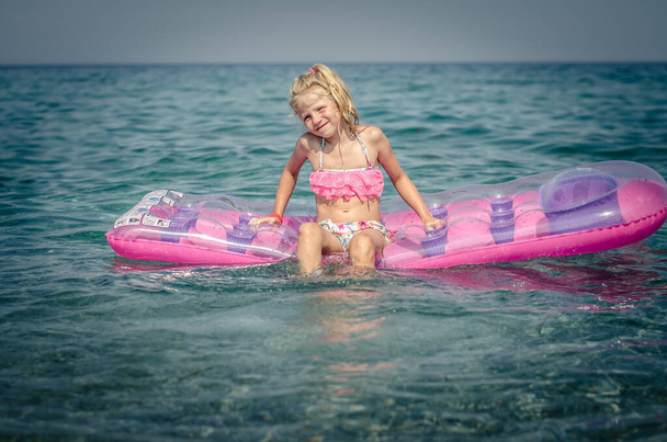 χαρούμενο χαμογελαστό παιδί με ξανθό κορίτσι που απολαμβάνει τις διακοπές του στη γαλάζια θάλασσα κάθεται σε ροζ στρώμα αέρα από καουτσούκ - Φωτογραφία, εικόνα