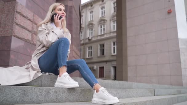 young attractive woman in a beige coat and jeans sitting on the stairs and talking on the phone on the street on the background of urban buildings. Sexy happy fashion girl smiling chatting smartphone - Video