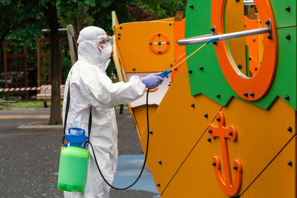 Children playground disinfecting and sanitizing against virus and disease. Man in white protective suit spraying disinfectant on slide to stop spreading coronavirus or COVID-19. Public health. - Photo, Image