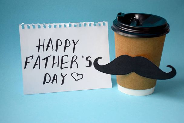 Happy Father`s Day card with cup of coffee decorated with paper mustache on blue background. - Photo, Image