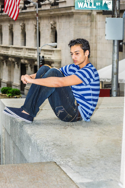 Magányos kelet-indiai amerikai tinédzser, aki kint gondolkodik New Yorkban, kék csíkos pólóban, törött farmerben, rongyos cipőben, Wall Street táblánál ül, karját ölelő lábakkal, szomorú - Fotó, kép