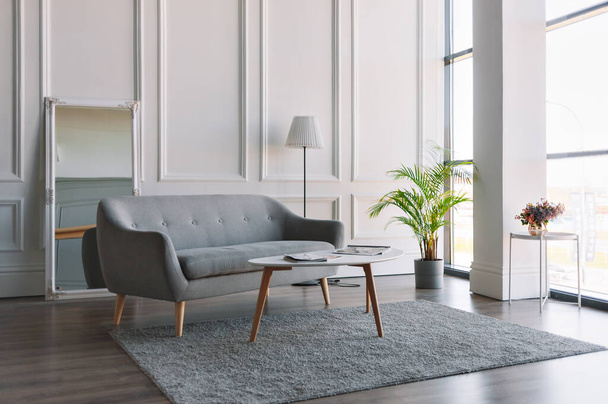 Grey and white interior in Scandinavian style. A grey sofa and a white table in a bright room with floor-length Windows. - Photo, Image