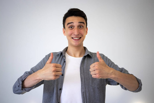 A cheerful, smiling guy gives a thumbs-up sign. On a gray background. A guy in a shirt and a white t-shirt shows the class with his fingers - Photo, image