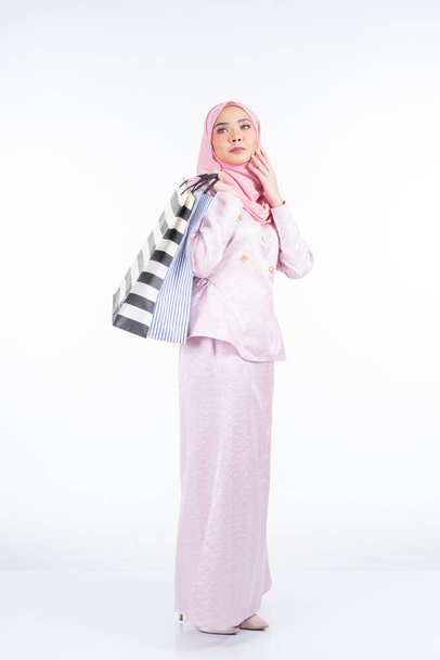 Ένα όμορφο μουσουλμανικό θηλυκό μοντέλο σε ένα ασιατικό παραδοσιακό φόρεμα μεταφέρουν τσάντες ψώνια που απομονώνονται σε λευκό φόντο. Idul fitri εορταστική προετοιμασία έννοια για ψώνια. Πλήρες μήκος πορτρέτο. - Φωτογραφία, εικόνα