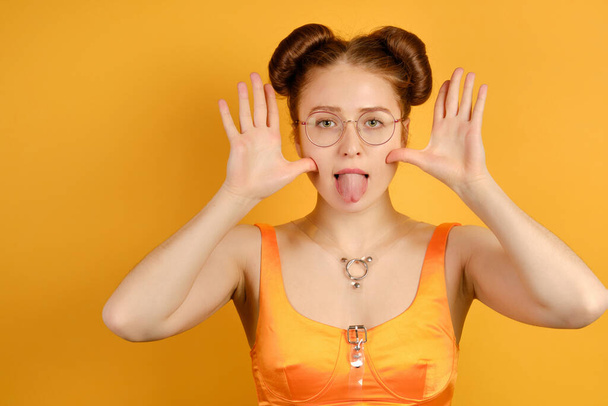 A red-haired girl in an orange top and round glasses stands on a yellow background, shows tongue, with open hands to face. - Foto, immagini