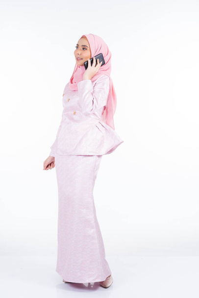 Μια ελκυστική μουσουλμάνα κυρία που φοράει παραδοσιακή ενδυμασία και χιτζάμπ, χρησιμοποιεί το smartphone της για selfie, chat και κάνοντας κλήση απομονωμένη σε λευκό φόντο - Φωτογραφία, εικόνα