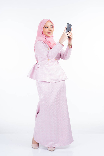 Μια ελκυστική μουσουλμάνα κυρία που φοράει παραδοσιακή ενδυμασία και χιτζάμπ, χρησιμοποιεί το smartphone της για selfie, chat και κάνοντας κλήση απομονωμένη σε λευκό φόντο - Φωτογραφία, εικόνα