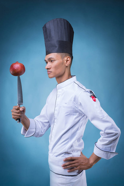 Een portret van een Aziatische chef-kok in zijn witte uniform en schort, zonder hoed op blauwe achtergrond spelend met zijn mes en een rode granaatappel - Foto, afbeelding